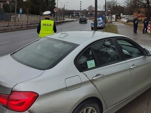 Na pierwszym planie stojący srebrny samochód, obok niego policjant ruchu drogowego, który stoi przy jezdni.