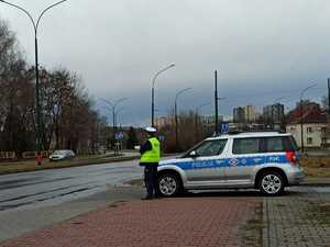 Policjant stojący przy radiowozie, który znajduje się prostopadle do drogi. Na drugim planie widoczne przejście dla pieszych.