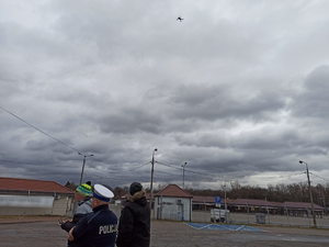 Policjant stojący obok dwóch mężczyzn. Jeden z nich kieruje dronem który znajduje się nad nimi.