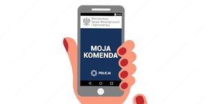 Grafika przedstawia dłoń, w której znajduje się telefon komórkowy. Na ekranie widoczny w górnej części orzeł, a obok niego napis Ministerstwo Spraw Wewnętrznych i Administracji, poniżej napis MOJA KOMENDA, a na dole znajduje się logo i napis POLICJA.