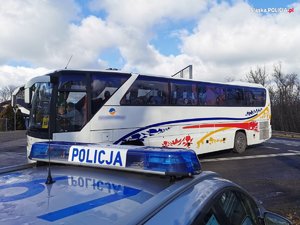 Na zdjęciu widać w oddali zaparkowany autokar koloru białego z kolorowymi naklejkami. Na pierwszym planie widać radiowóz policyjny. Zdjęcie zrobione w porze dziennej.