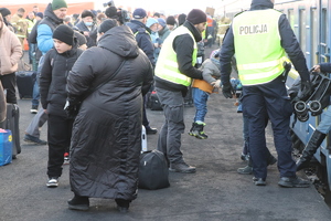 Po lewej stronie znajduje się grupa osób cywilnych, natomiast po prawej stronie widać dwóch policjantów, z czego jeden z nich trzyma dziecko na rękach.