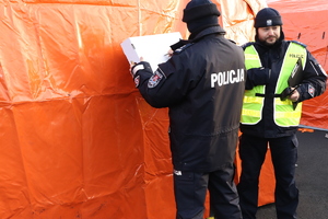 Zastępca Komendanta Powiatowego Policji w Będzinie mł.insp. Paweł Fryc, który na kartonie piszę nazwę miejscowości, aby ułatwić przyjezdnym kierowanie się do przygotowanych środków transportu. Obok niego znajduje się inny policjant.