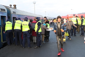 Grupa policjantów pomagająca przy wsiadaniu z pociągu. Na pierwszym planie widać uśmiechniętego młodego mężczyznę, który trzyma dwie bułki w ręce.