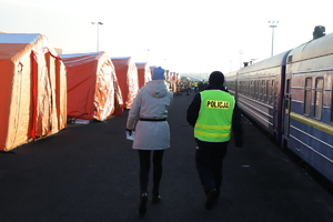 Policjant stojący z kobietą. Po prawej stronie znajduje się pociąg, natomiast po lewej stronie rozstawione pomarańczowe namioty.