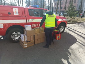 Policjant stojący przy samochodzie straży pożarnej, trzyma w ręku torbę ratowniczą, obok niego znajdują się kartony z zakupionymi rzeczami.