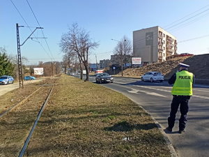 Policjant stojący z miernikiem prędkości przy krawędzi jezdni. Mierzy on prędkość jadących pojazdów.