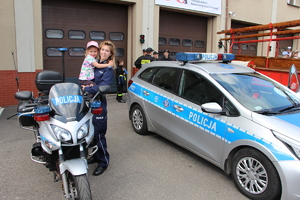 Policjantka trzymająca małą dziewczynkę na rękach pomiędzy motocyklem policyjnym i radiowozem.