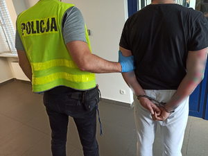 Policjant po cywilnemu, który ma założoną kamizelkę z napisem POLICJA trzymający zatrzymaną osobę.