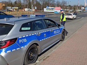 Na pierwszym planie radiowóz oraz policjant przy drodze. W oddali znajduje się przejście dla pieszych i przejeżdżające samochody.