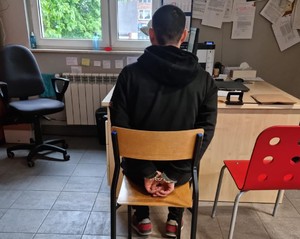 Zatrzymany mężczyzna, który ma założone kajdanki na ręce trzymane z tyłu, siedzący w jednym z pokoi na komisariacie.