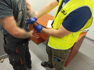 Policjant z założoną kamizelką odblaskową z napisem POLICJA zakuwa w kajdanki zatrzymanego