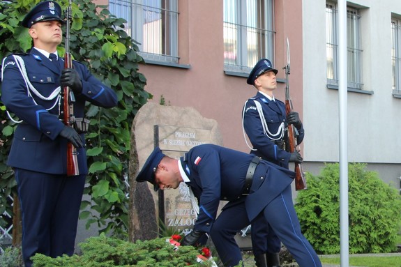 Policjant składa kwiaty nad obeliskiem, przy którym stoją policjanci tworząc wartę honorową.