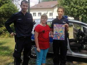 Policjanta i policjant stoją z dziewczynką przed radiowozem. Policjantka trzyma kolorową kartkę papieru.