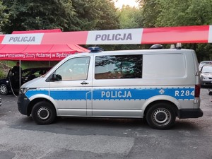 Na pierwszym planie widoczny policyjny radiowóz oraz taśma oddzielająca miejsce zdarzenia. Na drugim planie widoczny samochód pod namiotem oraz policjant wykonujący czynności.