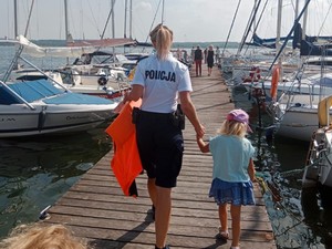 Policjantka trzyma za rękę dziewczynkę i idzie z nią przez pomost. Na drugim planie widoczne są łódki i inne osoby.