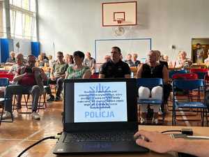 Zdjęcie przedstawia komputer z włączoną prezentacją przygotowaną przez policjantów, w tle widoczni zgromadzeni ludzie, którzy siedzą na krzesłach.