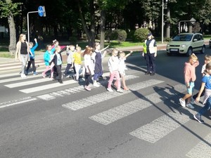 Dzieci przechodzą przez pasy na jezdni, obok pasów stoi umundurowana policjantka.