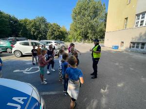 Dzieci stoją przed szkołą na parkingu i przemawia do nich policjantka.