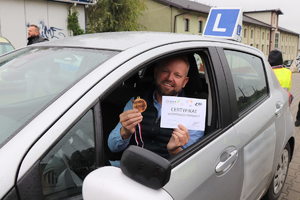 Kierowca samochodu do nauki jazdy trzyma otrzymany przez dzieci certyfikat wzorowego kierowcy oraz medal.