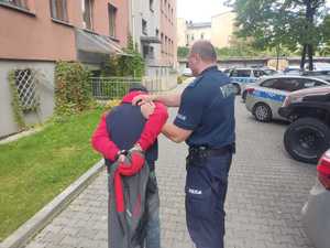 Policjant trzymający zatrzymanego, który ma założone kajdanki na ręce trzymane z tyłu na terenie parkingu będzińskiej komendy.