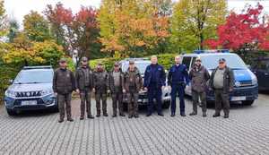 Strażnicy leśni wraz z policjantami stoją przed samochodami służbowymi.