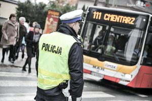 Na zdjęciu widać umundurowanego policjanta, który stoi na przejściu dla pieszych. W tle widać autobus z napisem cmentarz.