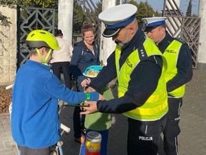 Policjanci w kamizelkach odblaskowych rozdają odblaski trzem chłopcom w kaskach. Na zdjęciu znajdują się jeszcze 2 kobiety.