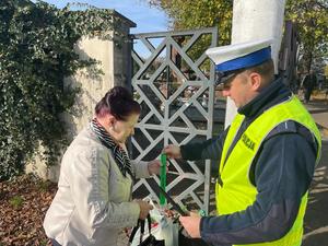 Policjant wręcza kobiecie opaskę odblaskową przed bramą cmentarza.