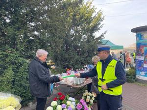 Policjant wręcza mężczyźnie sprzedającemu kwiaty przy cmentarzy odblask.