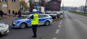 Policjant stoi przy dwóch radiowozach, które znajdują obok drogi, na drugim planie widać osoby sprzedające kwiatki, przechodniów oraz samochody.