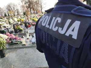Napis POLICJA na kamizelce policjanta, na drugim planie groby cmentarne.