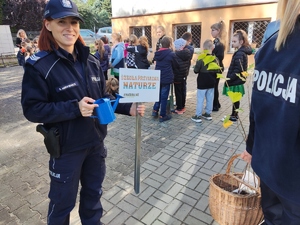 Policjantka trzyma w ręce tabliczkę &quot;Szkołą przyjazna naturze&quot; oraz konewkę, za nią znajdują się dzieci na placu przed szkołą.