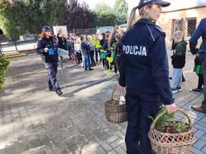 Dwie policjantki na pierwszym planie, jedna trzyma w ręce tabliczkę i konewkę, druga kosze z drzewkami do sadzenia. Za nimi znajduje się grupa dzieci z wychowawcą.