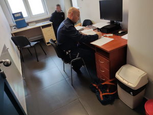 Policjant siedzi przy biurku i wykonuje czynności, na końcu pomieszczenia siedzi mężczyzna, który ma założone kajdanki na ręce trzymane z tyłu.