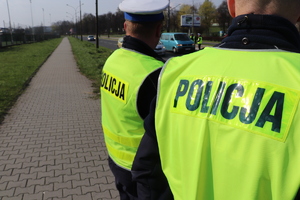 Na pierwszym planie zbliżenie na dwóch policjantów, którzy mają założone kamizelki odblaskowe z napisem POLICJA. na drugim planie policjanci na drodze sprawdzają trzeźwość kierujących za pomocą alkomatu.