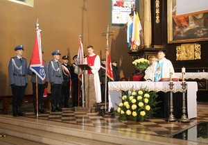 Zdjęcie przedstawia 3 policjantów, którzy tworzą sztandar Komendy Powiatowej Policji w Będzinie, ustawiony w kościele przy ołtarzu. Obok nich znajduje się inny mundurowy sztandar oraz czytający ministrant, a także księża stojący przy ołtarzu.