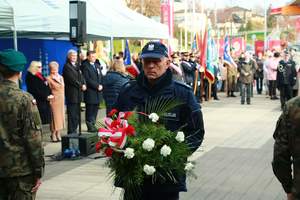 Policjant trzymający kwiaty do złożenia, za nim grupa osób podczas uroczystych Obchodów Dnia Niepodległości.