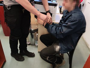 Na zdjęciu policjant zakładający kajdanki zatrzymanemu mężczyźnie, który siedzi na krześle.