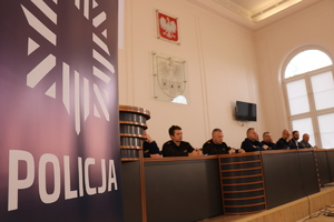 Na zdjęciu po lewej stronie na niebieskim tle napis POLICJA w oddali widać osoby przemawiające podczas debaty.