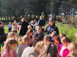 Zdjęcie przedstawia policjantkę i grupę dzieci zebranych wokół przed sadzeniem drzewka.