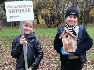 Na zdjęciu dwie dziewczynki. Jedna trzyma w ręce tabliczkę z napisem&quot; Szkoła przyjazna naturze&quot;, druga trzyma drewniany domek dla ptaków.
