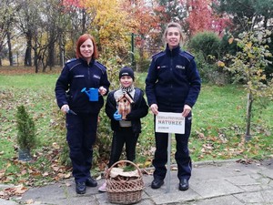 Zdjęcie przedstawia dwie policjantki i dziewczynkę. Trzymają one narzędzia do sadzenia, drewniany domek dla ptaków oraz tabliczkę informacyjną.