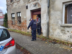 Na zdjęci policjant stoi przed budynkiem, z którego wychodzi mężczyzna.