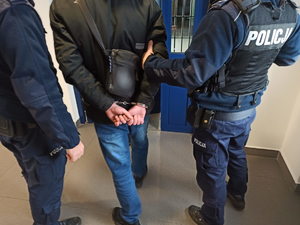 Na zdjęciu zatrzymany mężczyzna stoi z założonymi kajdankami na ręce obok umundurowanych policjantów.