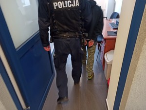 Na zdjęciu policjant wprowadza zatrzymanego, który ma założone kajdanki do pokoju.