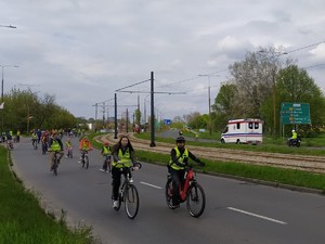 Na zdjęciu z przodu widoczni rowerzyści jadący w kamizelkach odblaskowych po ulicy, z tyłu widoczny policjant na motorze jadący za karetką pogotowia.