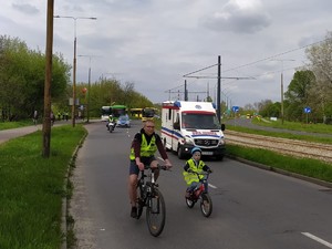 Na zdjęciu z przodu jedzie na rowerze chłopiec oraz mężna, za nim porusza się karetka pogotowia oraz policjant na motorze i radiowóz policyjny oraz autobusy.