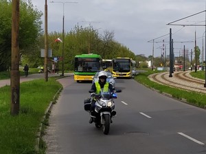 Na zdjęciu z przodu jedzie policjant na motorze, za nim radiowóz oraz autobusy.