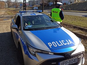 Na zdjęciu znajduje się radiowóz policyjny, a obok niego umundurowany policjant.